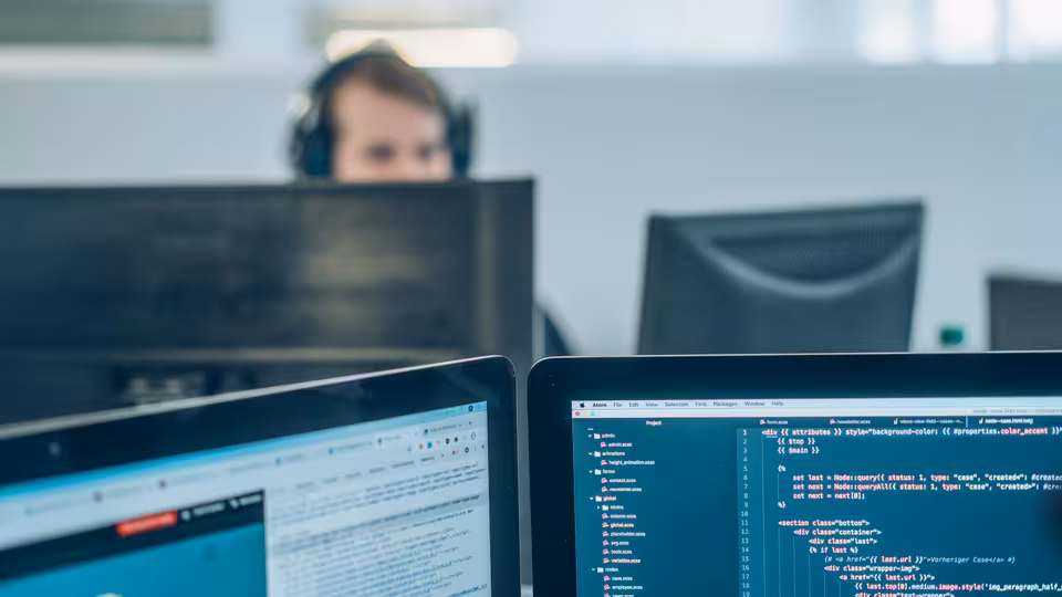 Mehrere Computerbildschirme, im Hintergrund eine Person