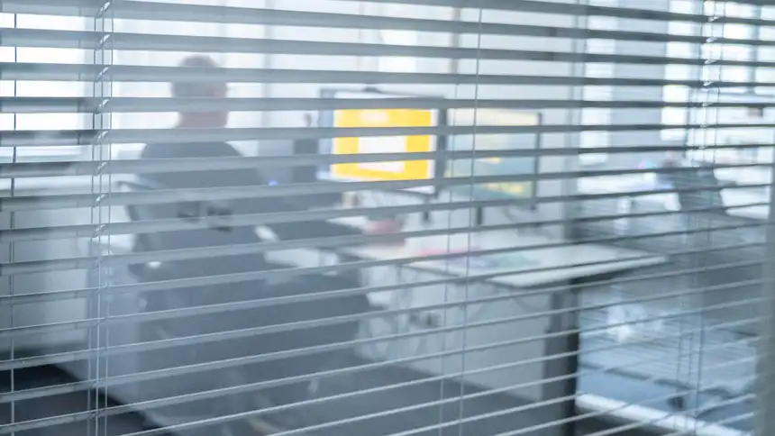 Eine Person sitzt an einem Schreibtisch und schaut auf den Monitor. Im Vordergrund sieht man verschwommen eine Jalousie.