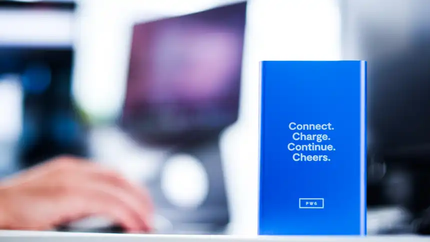 Eine Powerbank mit einem Slogan, im Hintergrund eine Person, die am Schreibtisch sitzt.