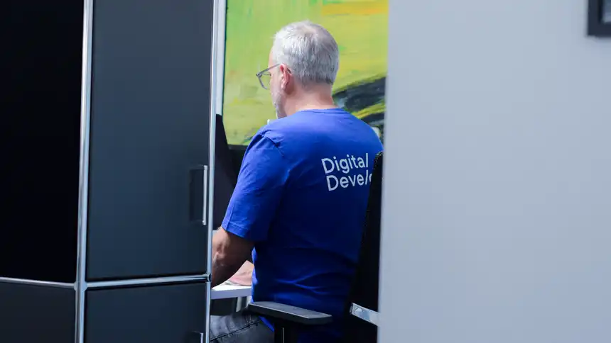 aEine Person mit einem blauen T-Shirt mit der Aufschrift "Digital Developers" sitzt vor einem Monitor 