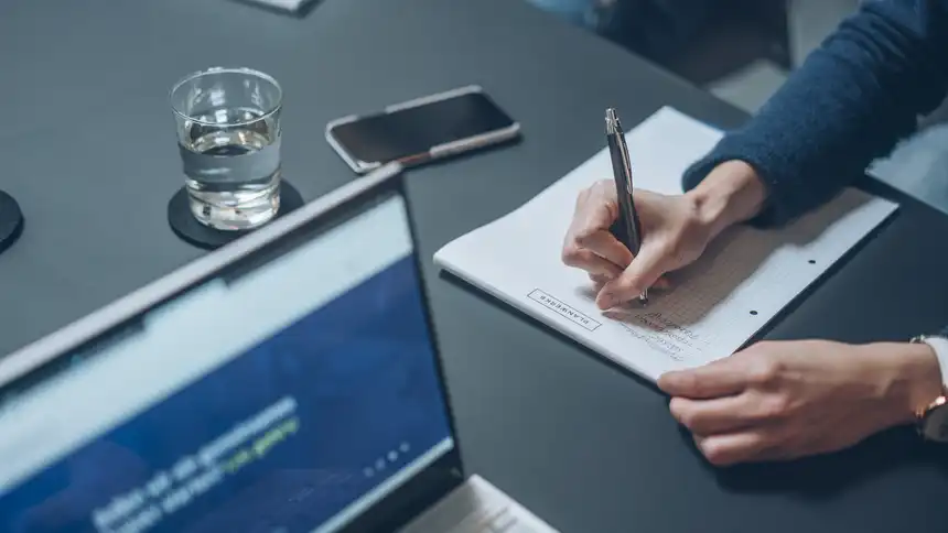 Eine Person schreibt etwas auf einen Block, im Vordergrund ist ein Laptop zu sehen