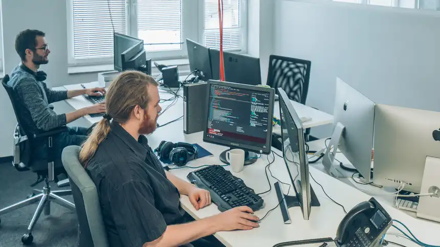 Zwei Personen sitzen an Schreibtischen vor einem Monitor und Tastatur