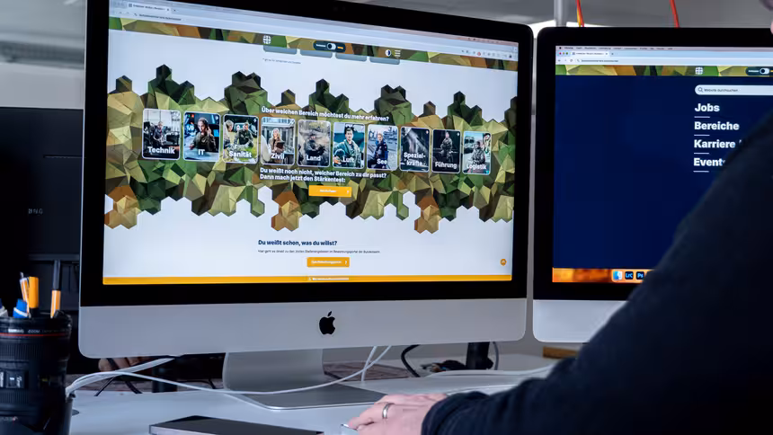 Zwei Apple Monitore, die die Karriereseite der Bundeswehr zeigen, davor eine Person, die auf den Bildschirm schaut.