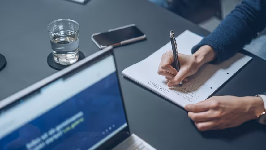 Eine Person schreibt etwas auf einen Block, im Vordergrund ist ein Laptop zu sehen