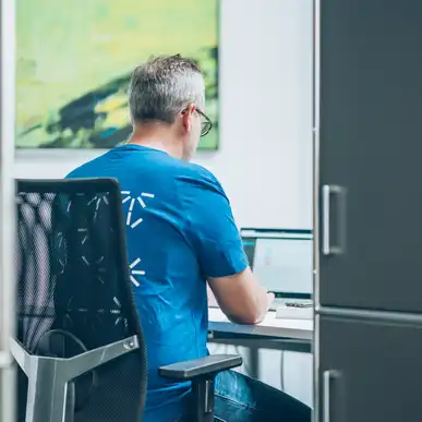 Eine Person in blauem T-Shirt sitzt am Schreibtisch vor einem Laptop